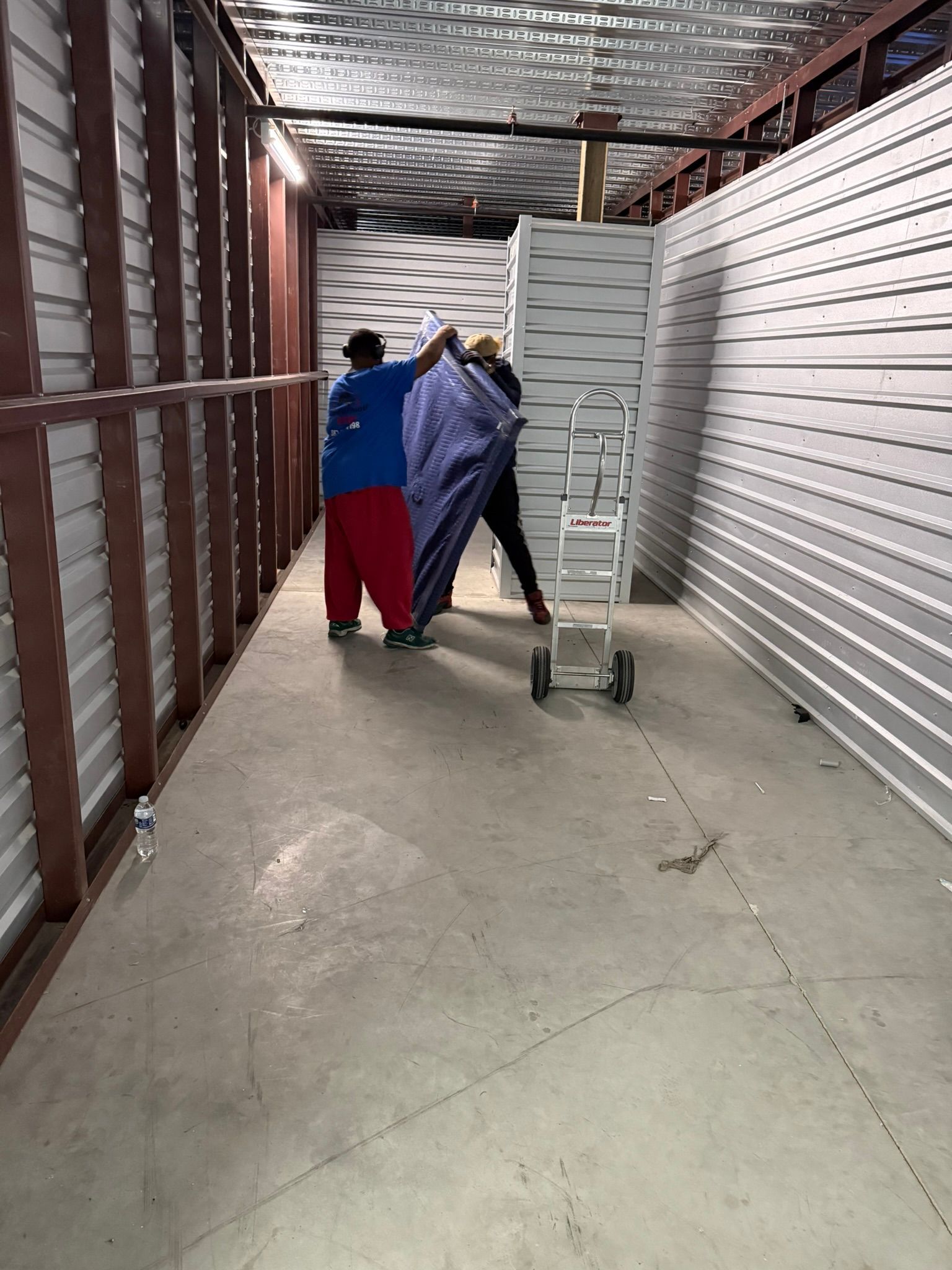 Two people loading an item wrapped in blue material into a storage unit with a dolly.