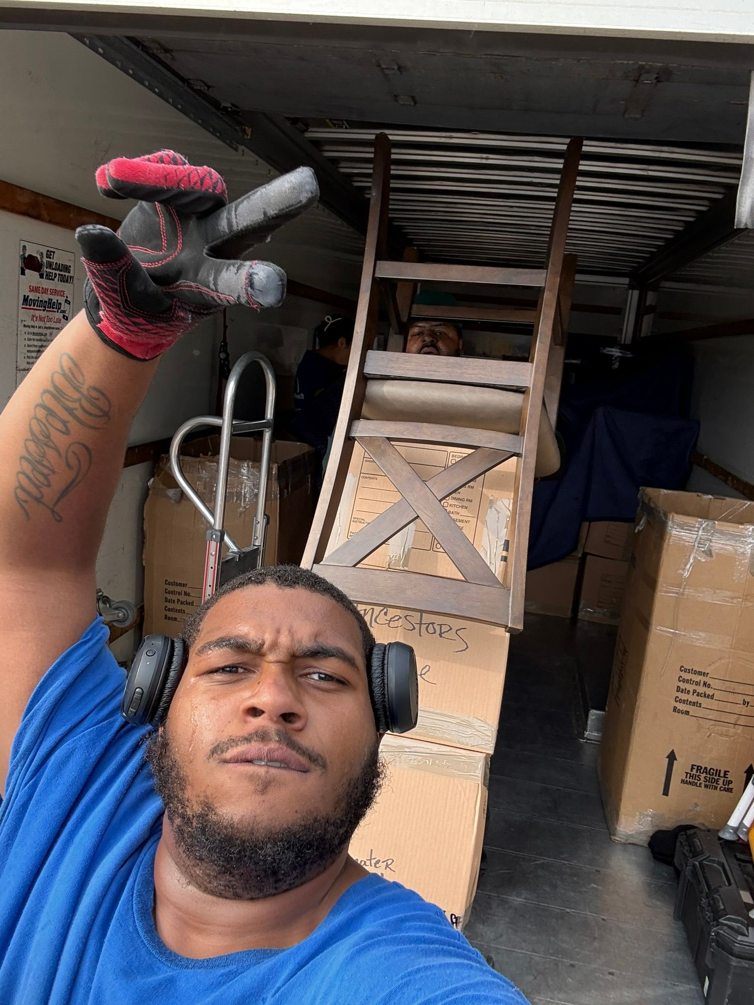 Man in blue shirt, gesturing inside a moving truck with boxes and furniture. Another person on a raised platform.