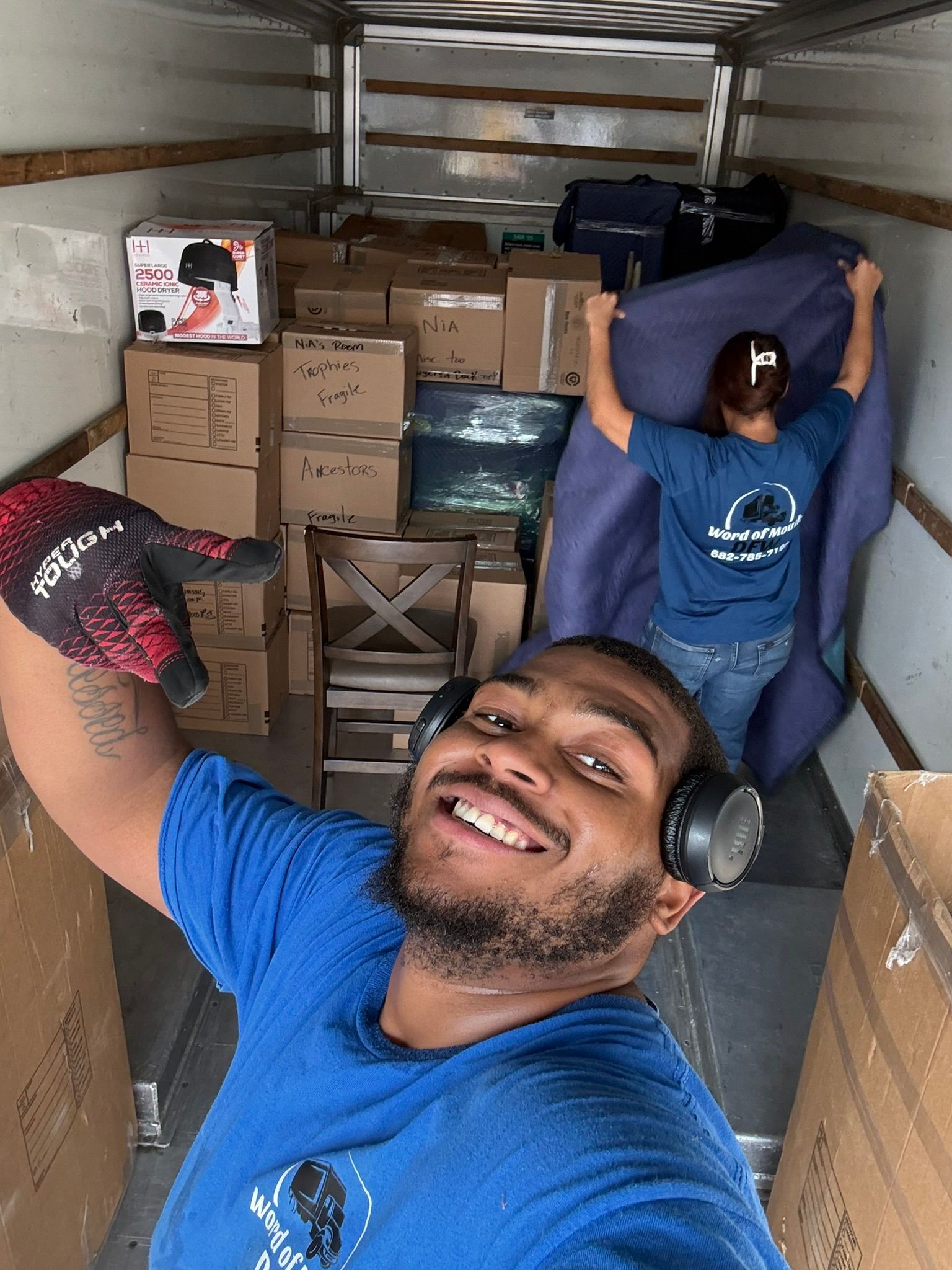 Two movers in truck; man smiles at camera, other holds blanket. Boxes and furniture are loaded.
