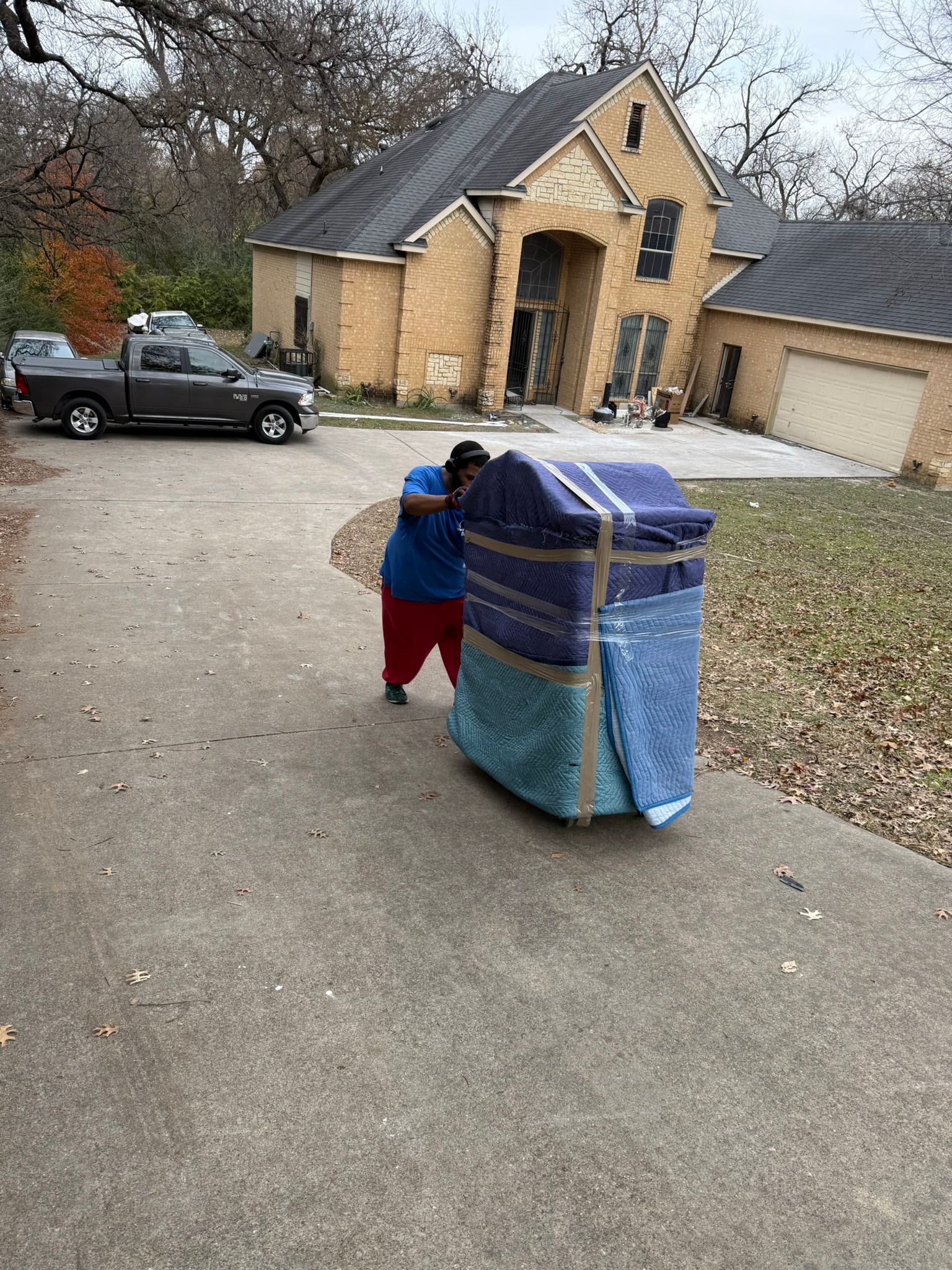 A person carrying a large, wrapped object up a driveway toward a house with a truck parked nearby.