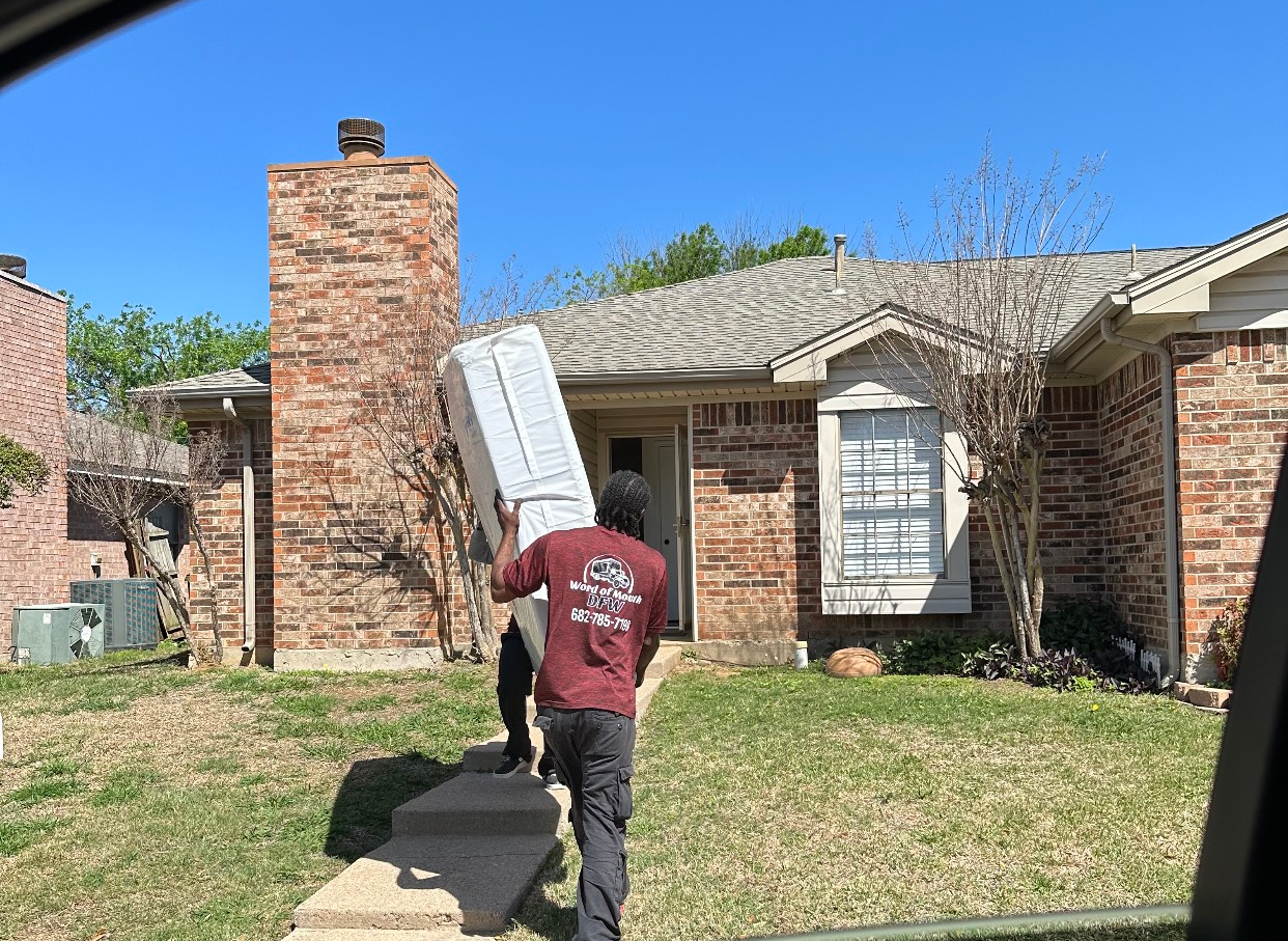 Two movers carrying a large, white appliance into a brick house on a sunny day.
