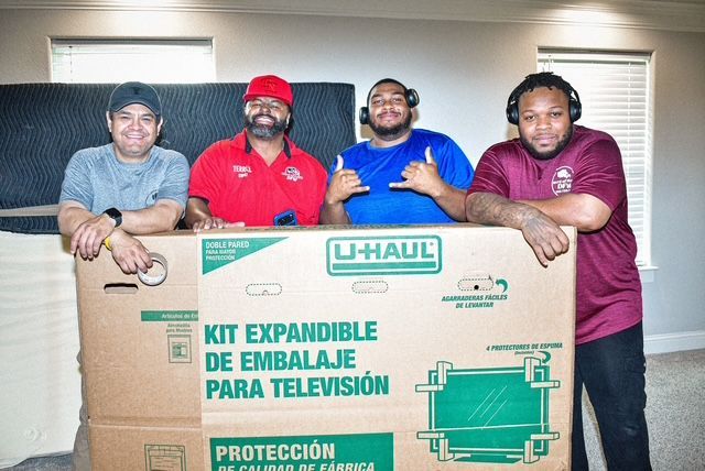 Four people pose behind a large U-Haul TV packing kit box indoors.