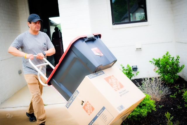 Person pushing a dolly loaded with boxes and a bin outside a house