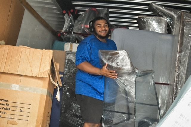 Man in blue shirt moving wrapped furniture in a storage area filled with boxes and packing materials