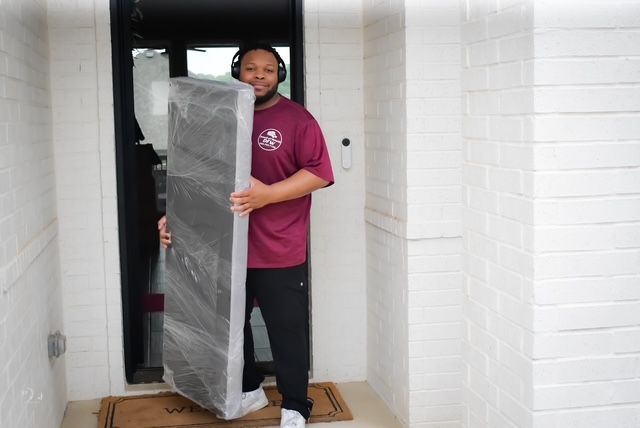 Person carrying a tall wrapped item through a doorway in a white hallway.