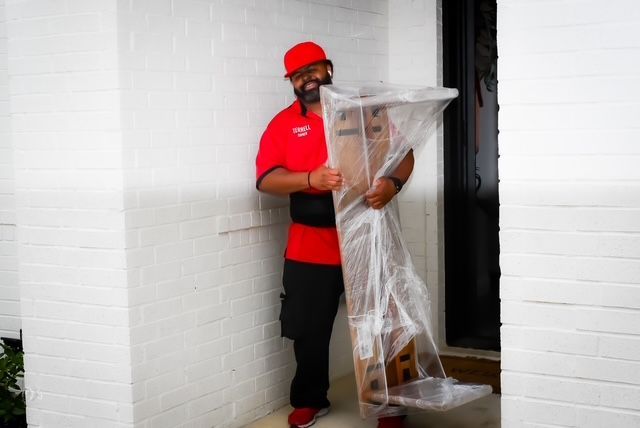 Man in red cap and shirt carrying a wrapped mirror or frame in a doorway
