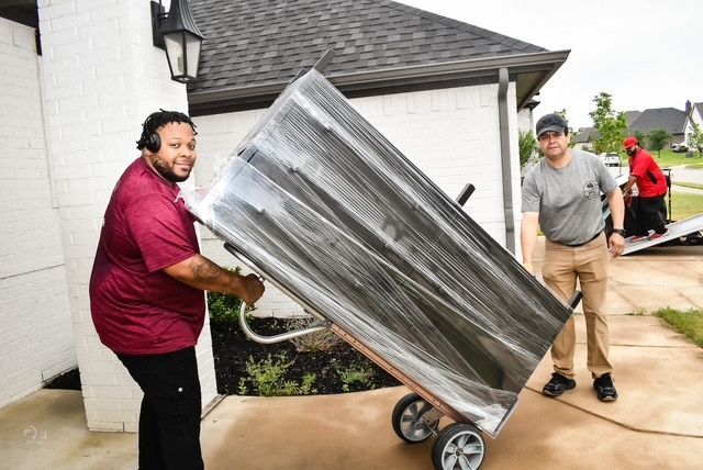 Two people moving a large wrapped item on a hand truck outside a house.