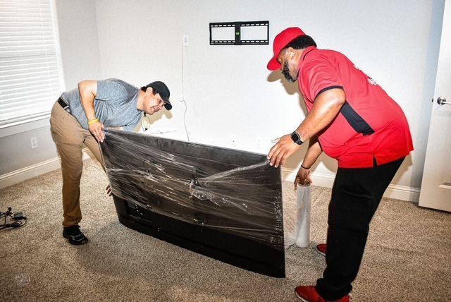 Two movers wrapping a TV in plastic in a carpeted room