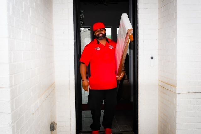 Man in red shirt standing in a doorway holding a white board, in a narrow white hallway