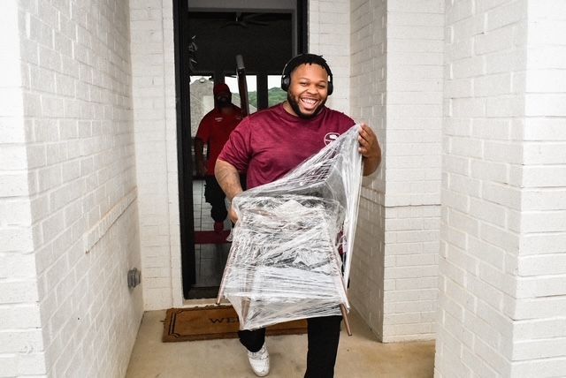 Man carrying a plastic-wrapped chair through a doorway, smiling in a narrow entryway