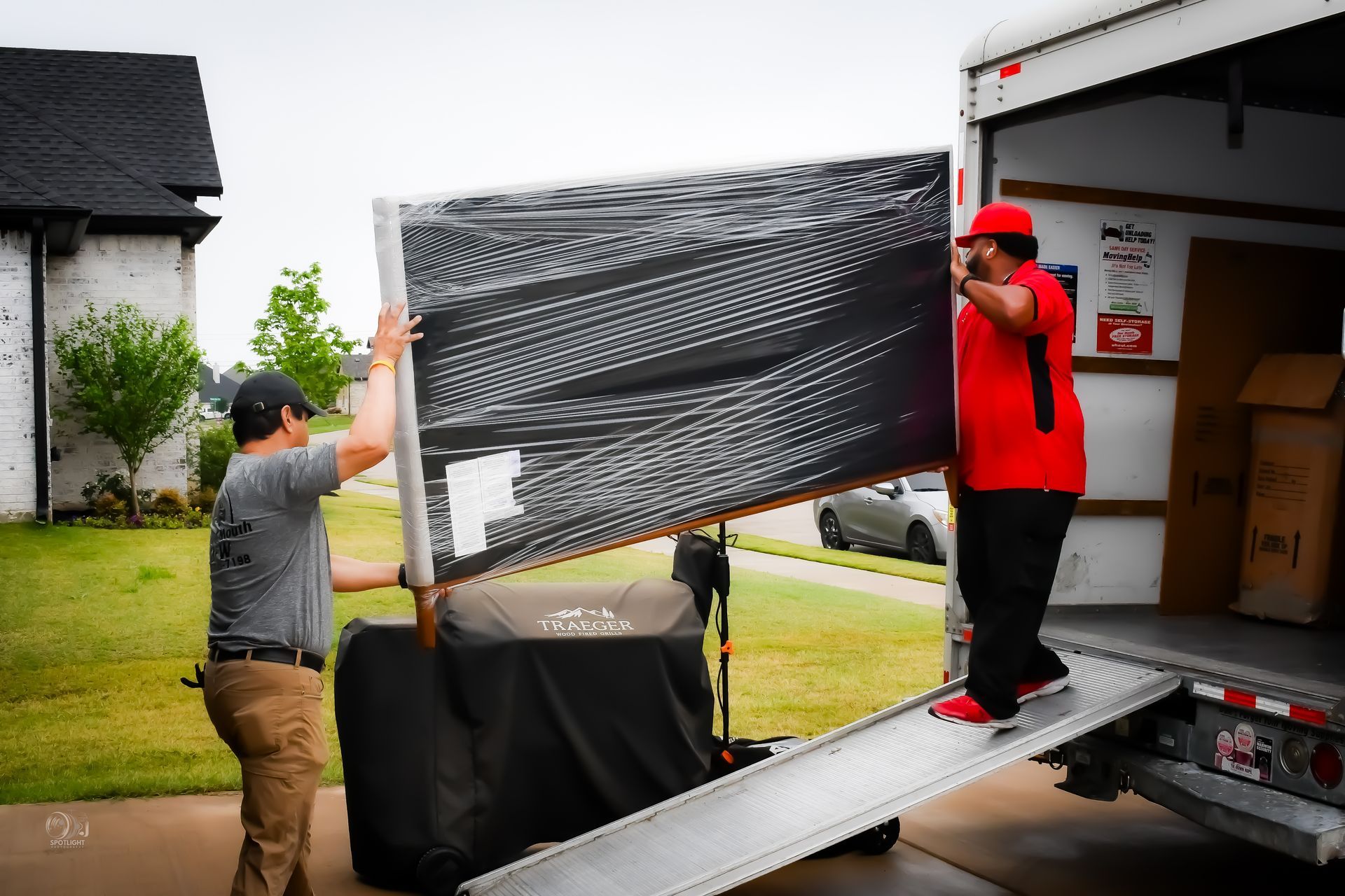Two people move a wrapped black item from a moving truck onto a dolly.