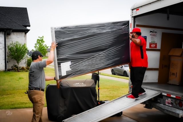 Two movers load a large wrapped TV into a truck from a driveway