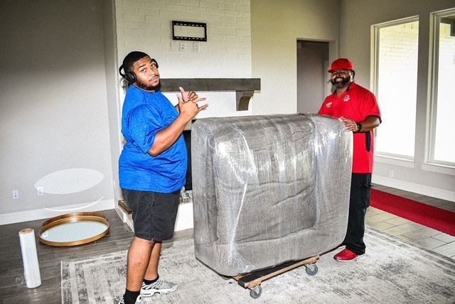 Two people moving a gray wrapped couch in a living room