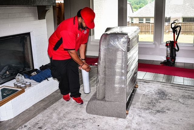 Worker in red uniform wrapping a chair in protective plastic inside a living room.