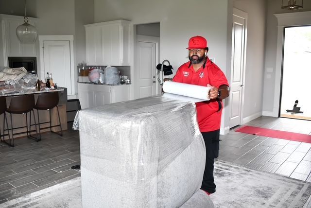 Mover in red shirt and hard hat carrying a wrapped sofa in a bright living room