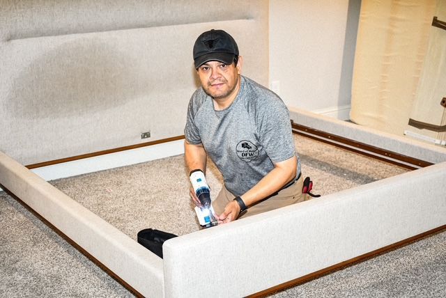 Man assembling a beige upholstered bed frame with a power drill in a carpeted bedroom