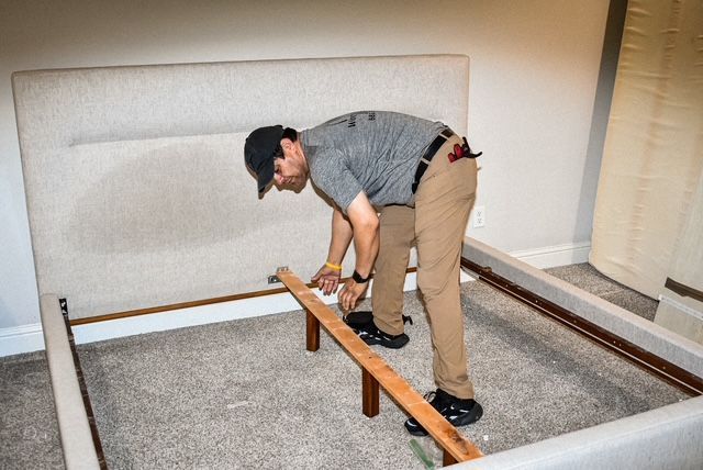 Person assembling a bed frame with a tool in a bedroom, leaning over the frame.