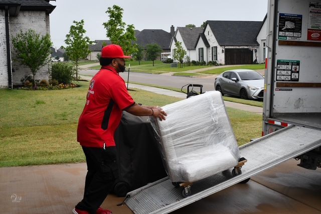 Delivery worker in red uniform pushing a pallet of wrapped boxes up a truck ramp.