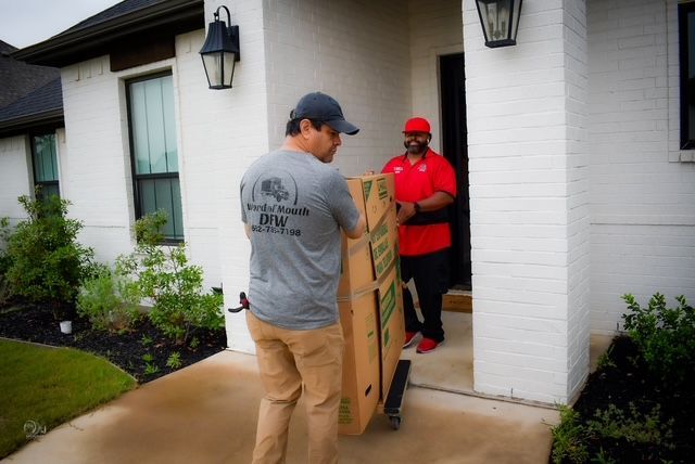 Two movers carrying a tall wooden cabinet into a white house through the front door