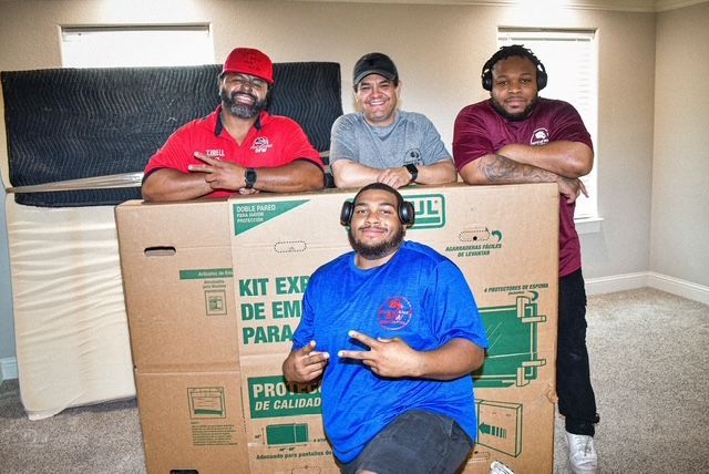 Four people posing behind and in front of a cardboard box, smiling in a room with beige walls
