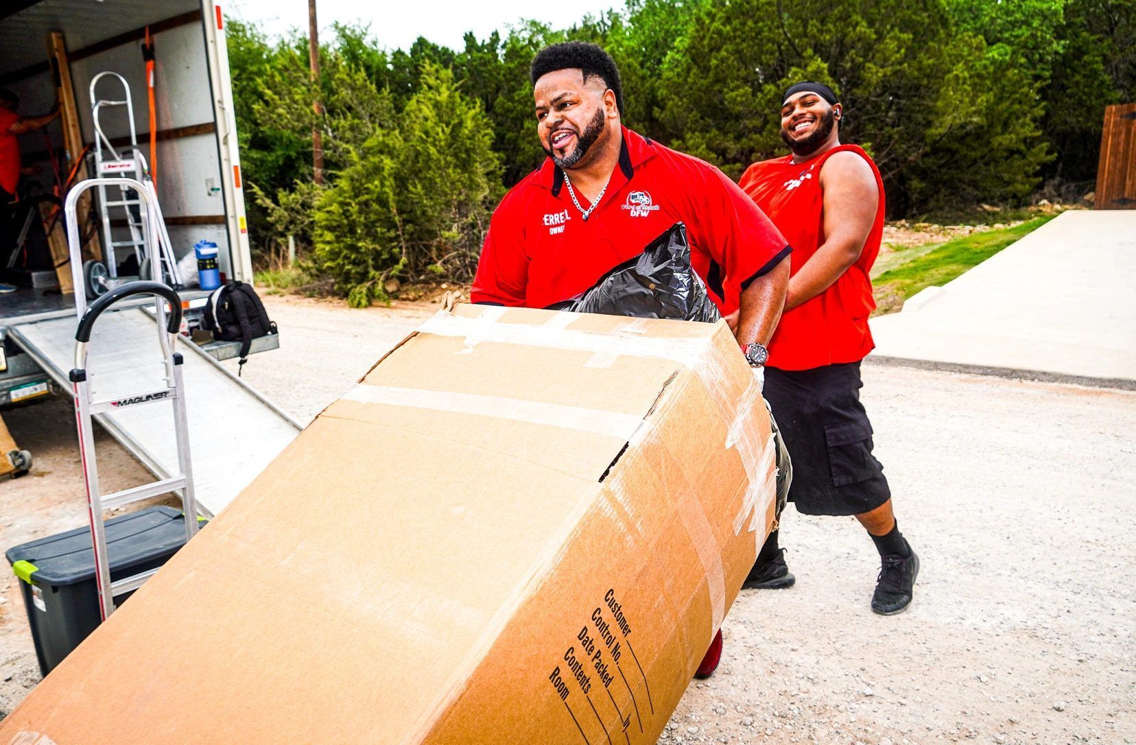Two men in red shirts are carrying a large cardboard box.