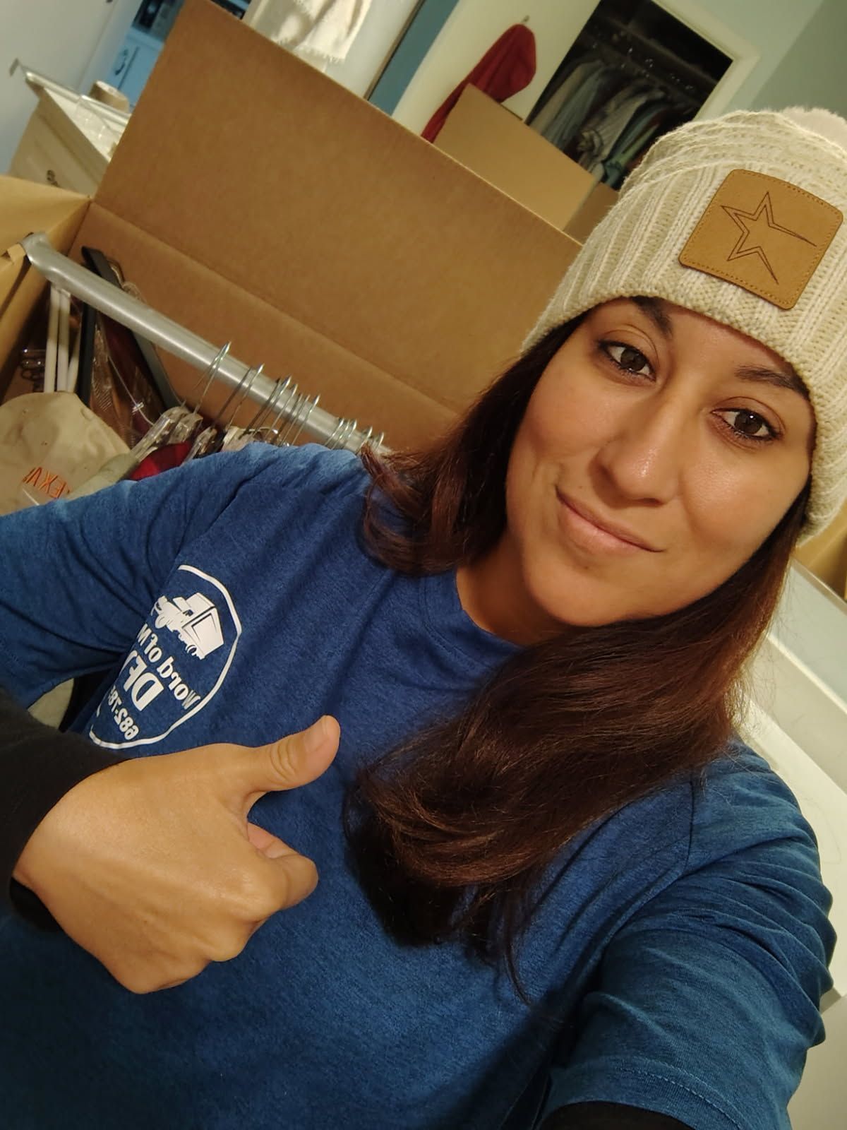 Person wearing a beanie and blue shirt giving a thumbs-up. Indoors, cardboard boxes and clothing racks in the background.