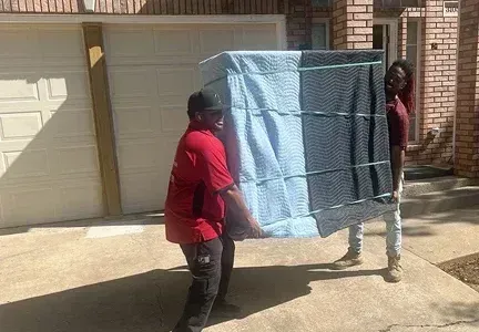 Two men are carrying a large piece of furniture in front of a garage.