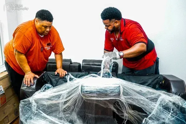 Two men are standing next to a couch wrapped in plastic.