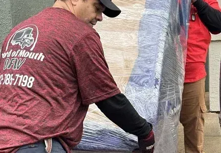 A man in a red shirt is carrying a couch wrapped in plastic.