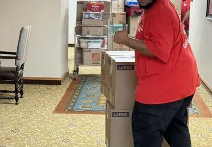 A man in a red shirt is carrying a large cardboard box.