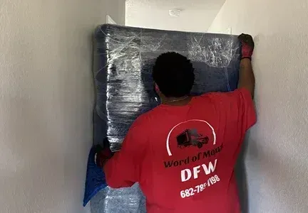A man in a red shirt is wrapping a mattress in plastic.
