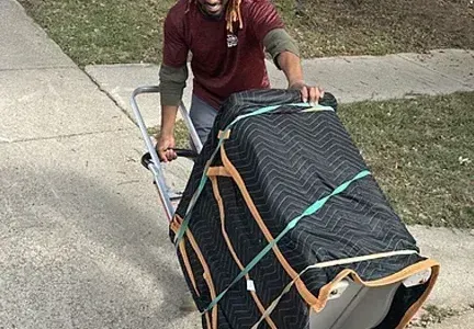 A man is pushing an appliance on a dolly down a sidewalk.