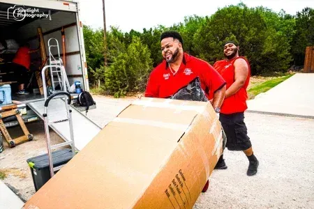 Two men in red shirts are carrying a large cardboard box.