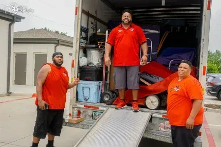 Three men are standing on the back of a moving truck.