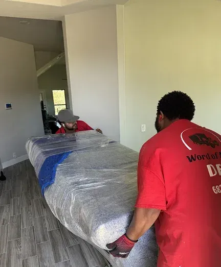 A man in a red shirt is carrying a mattress in a room.