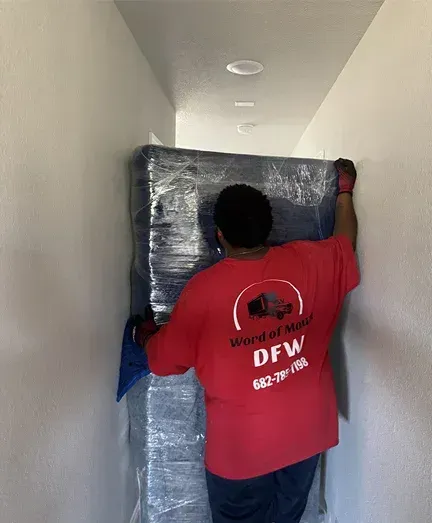A man in a red shirt is carrying a mattress wrapped in plastic.