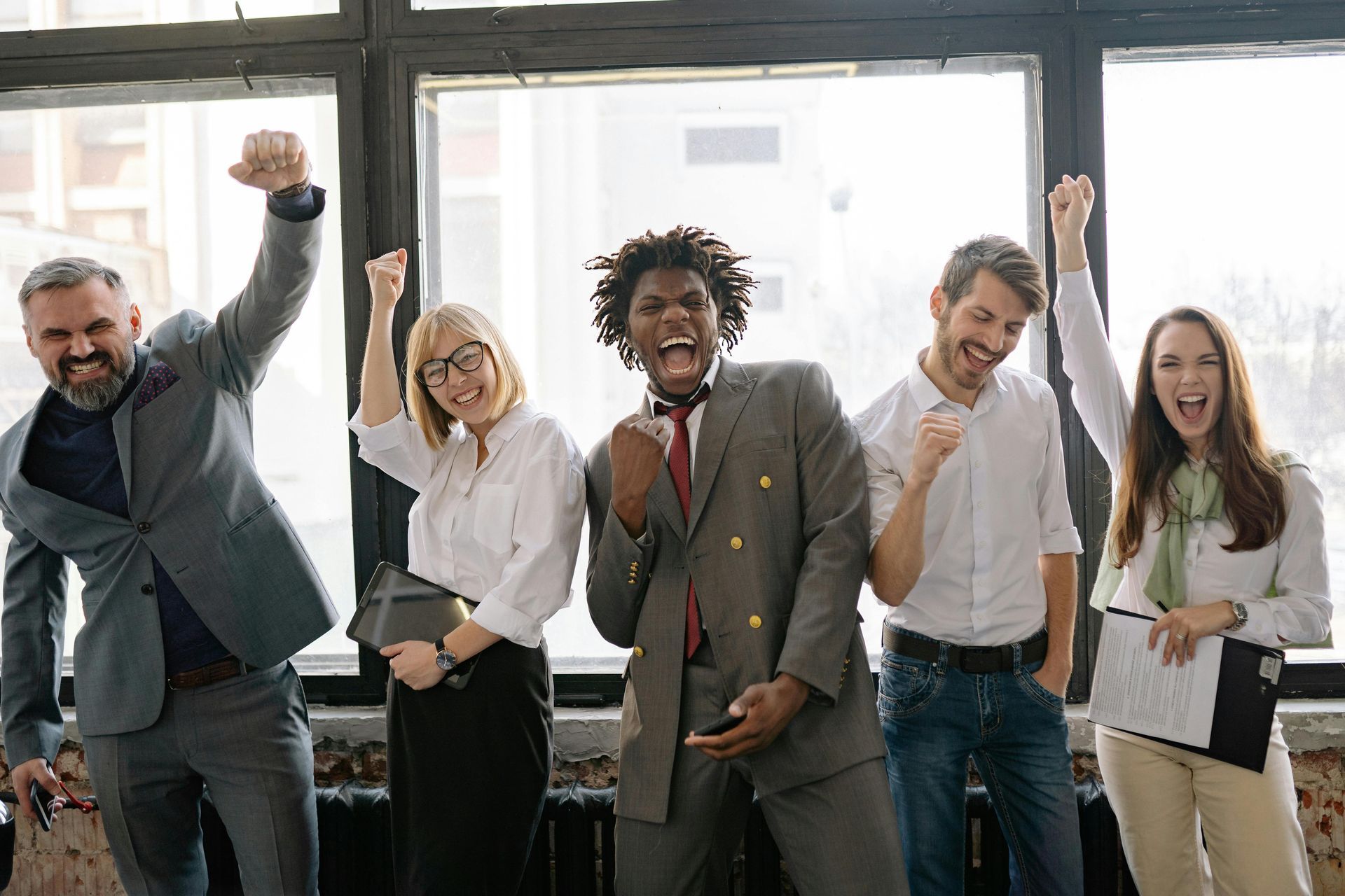 Group of diverse office workers celebrating with raised fists and excited expressions in front of a large window.