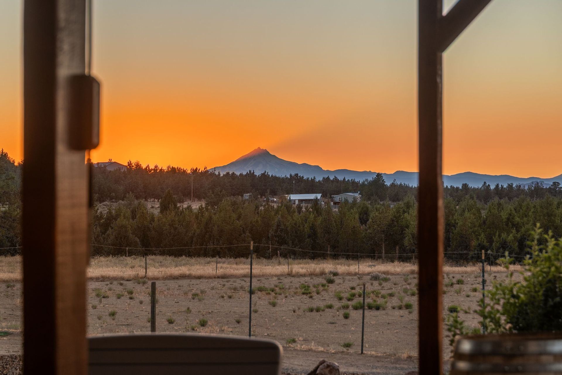 Sunset view through a wooden structure, mountain in the distance, orange sky, trees in between.