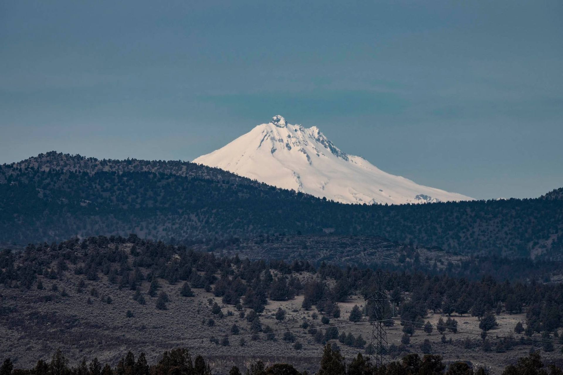 A snowy mountain with trees in the foreground and a blue sky in the background.