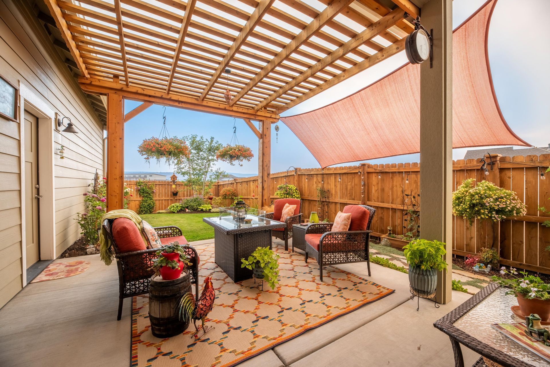 A patio with a wooden pergola, seating around a fire pit, and a shade sail above, set against a wooden privacy fence.