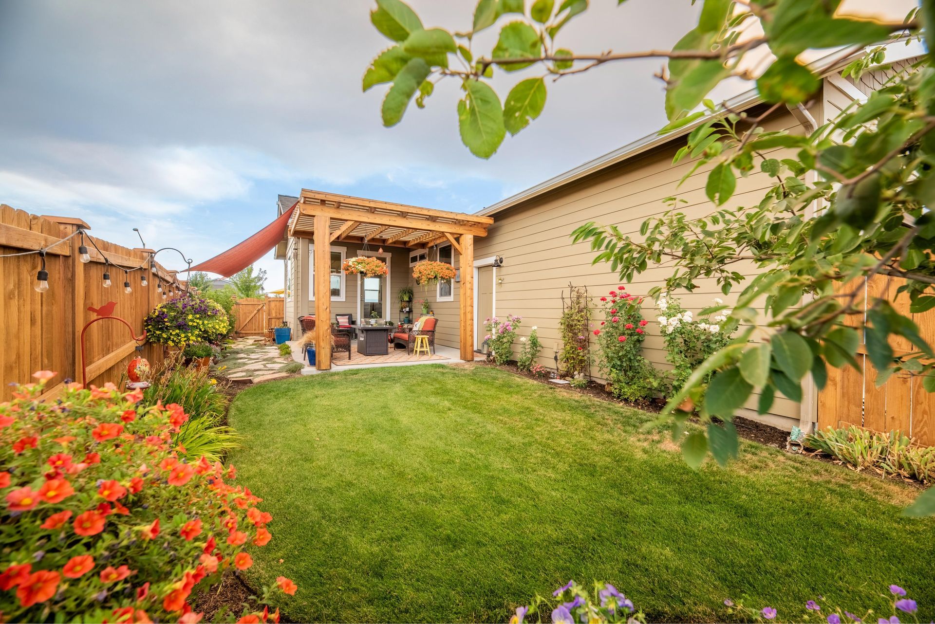 A lush backyard with a green lawn, orange flowers in the foreground, and a wooden pergola covering a patio seating area.