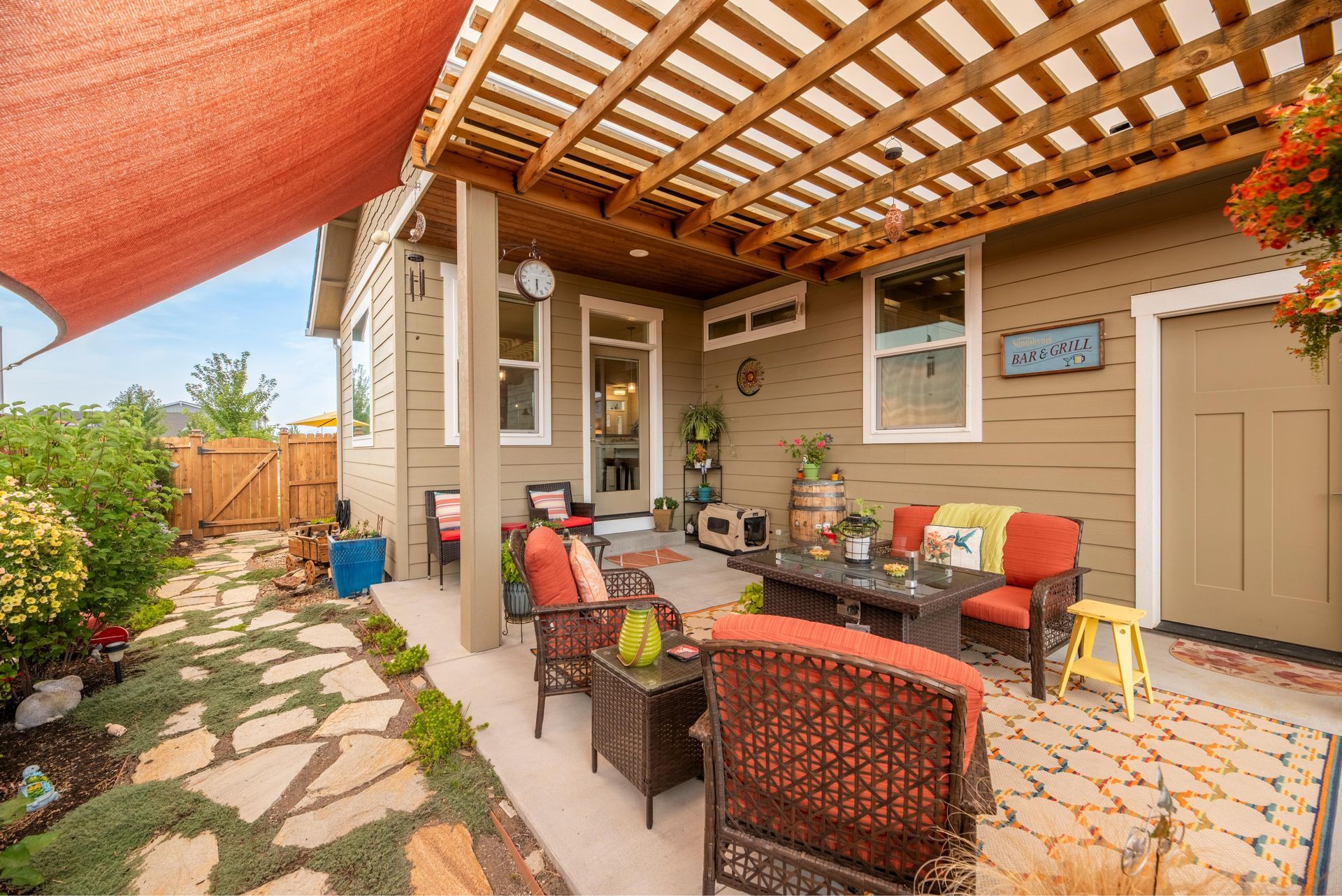 A patio area with a wooden pergola, outdoor seating, a stone walkway, and a red shade sail over a tan house exterior.