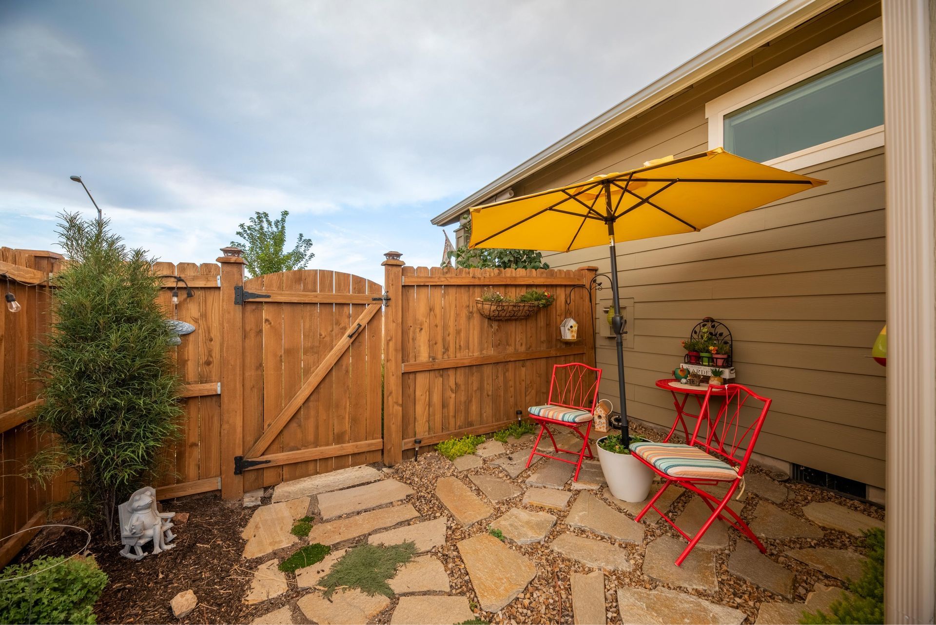 A small stone patio features two red chairs under a bright yellow umbrella, nestled next to a house and wooden fence.