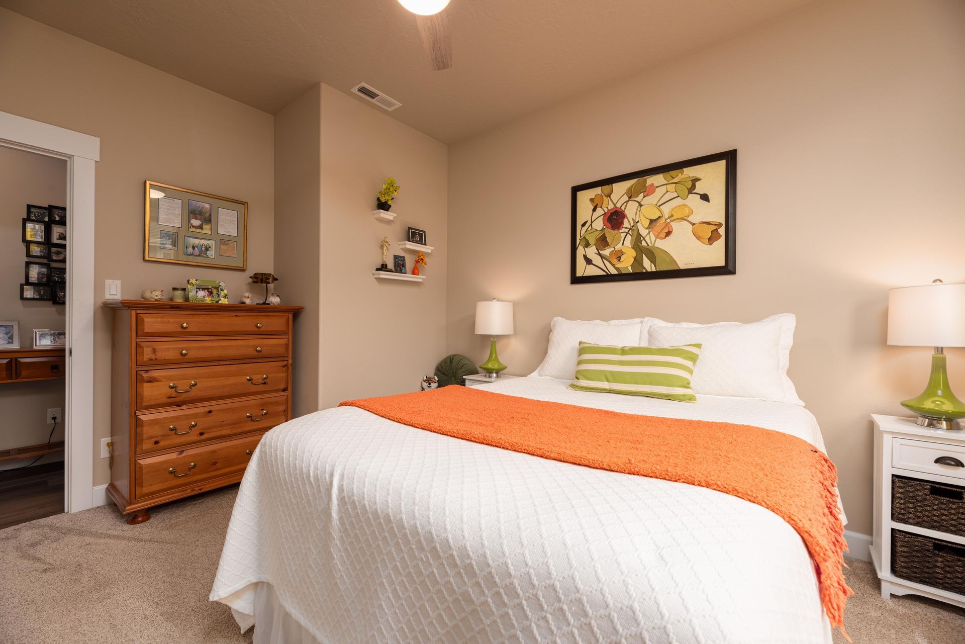 A bedroom with a queen bed featuring a white quilt and orange throw, a wooden dresser, and white nightstands with lamps.