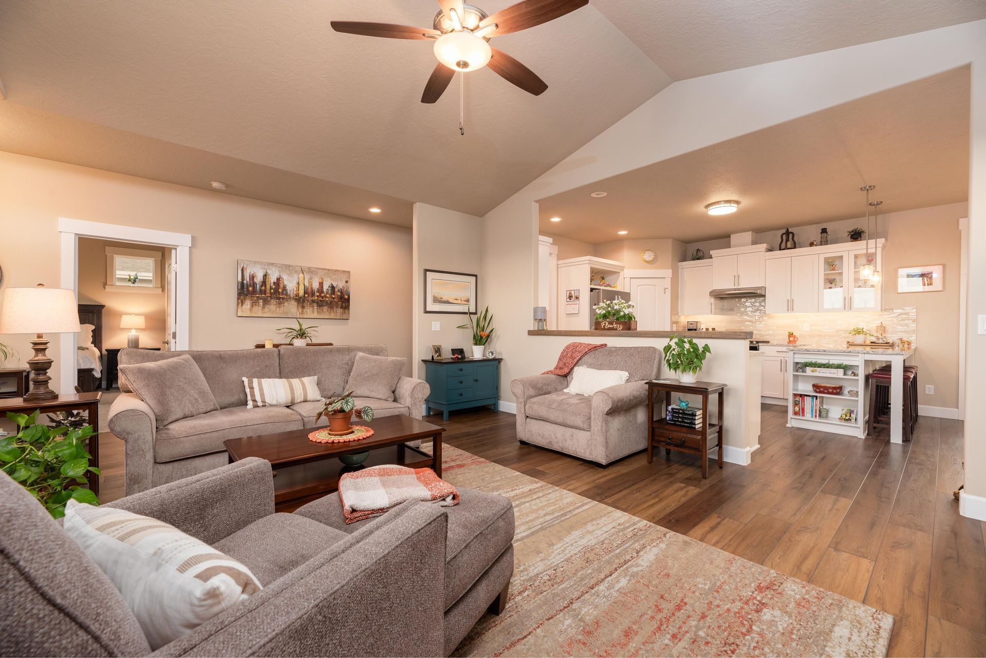 Living room and open kitchen with light brown wood flooring, grey furniture, vaulted ceilings, and a ceiling fan.