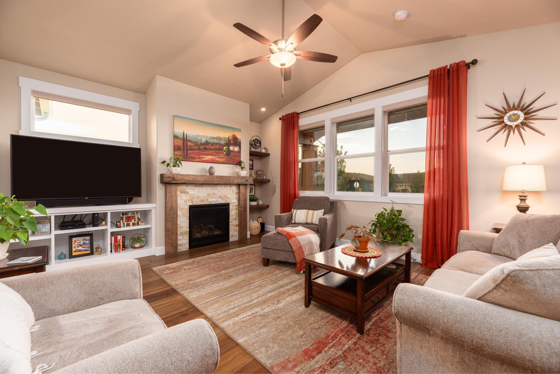 Cozy living room with a stone fireplace, large television, beige seating, wood floors, and orange curtains.