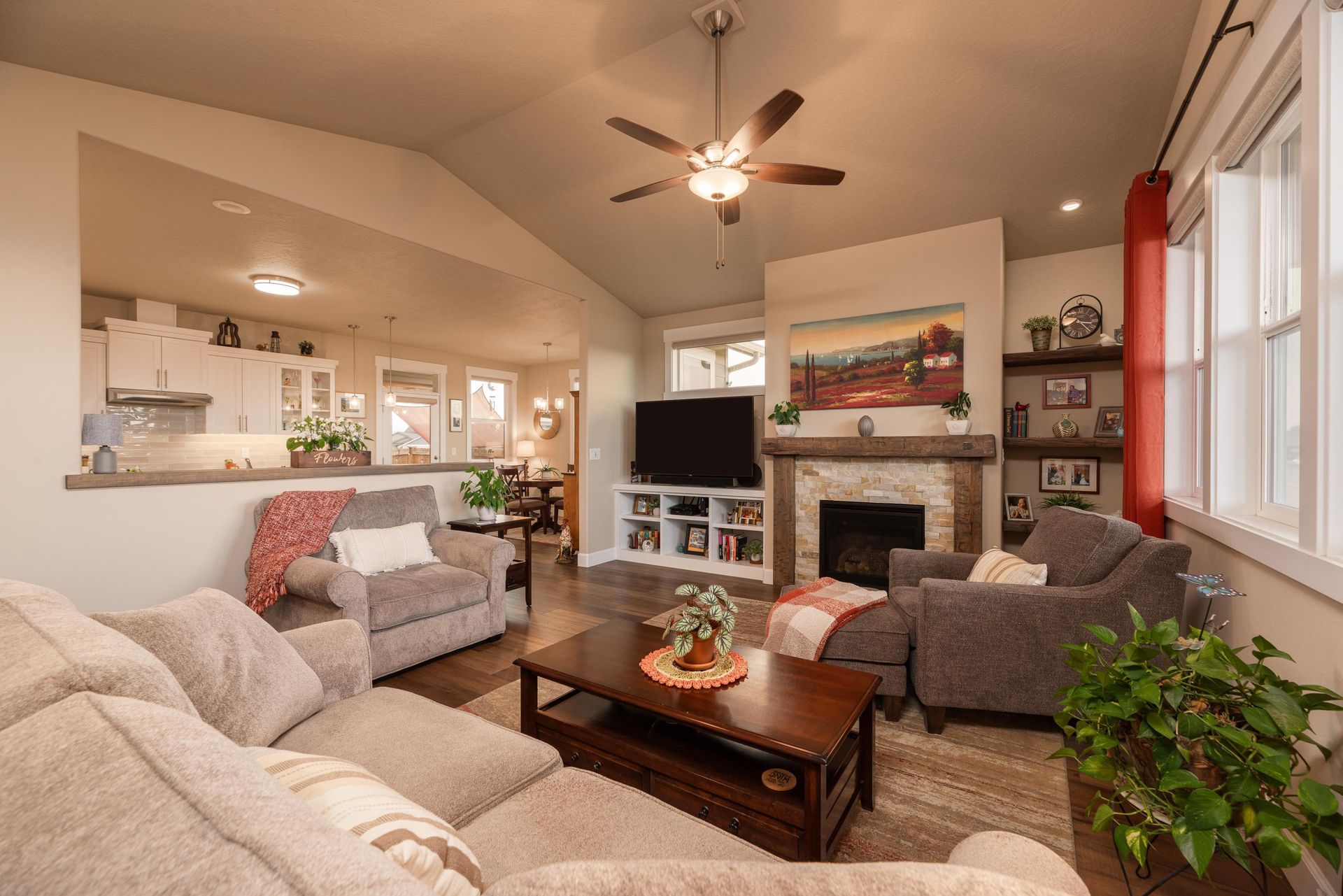 A cozy living room with grey seating, a stone fireplace, a wooden coffee table, and an open view into the kitchen.