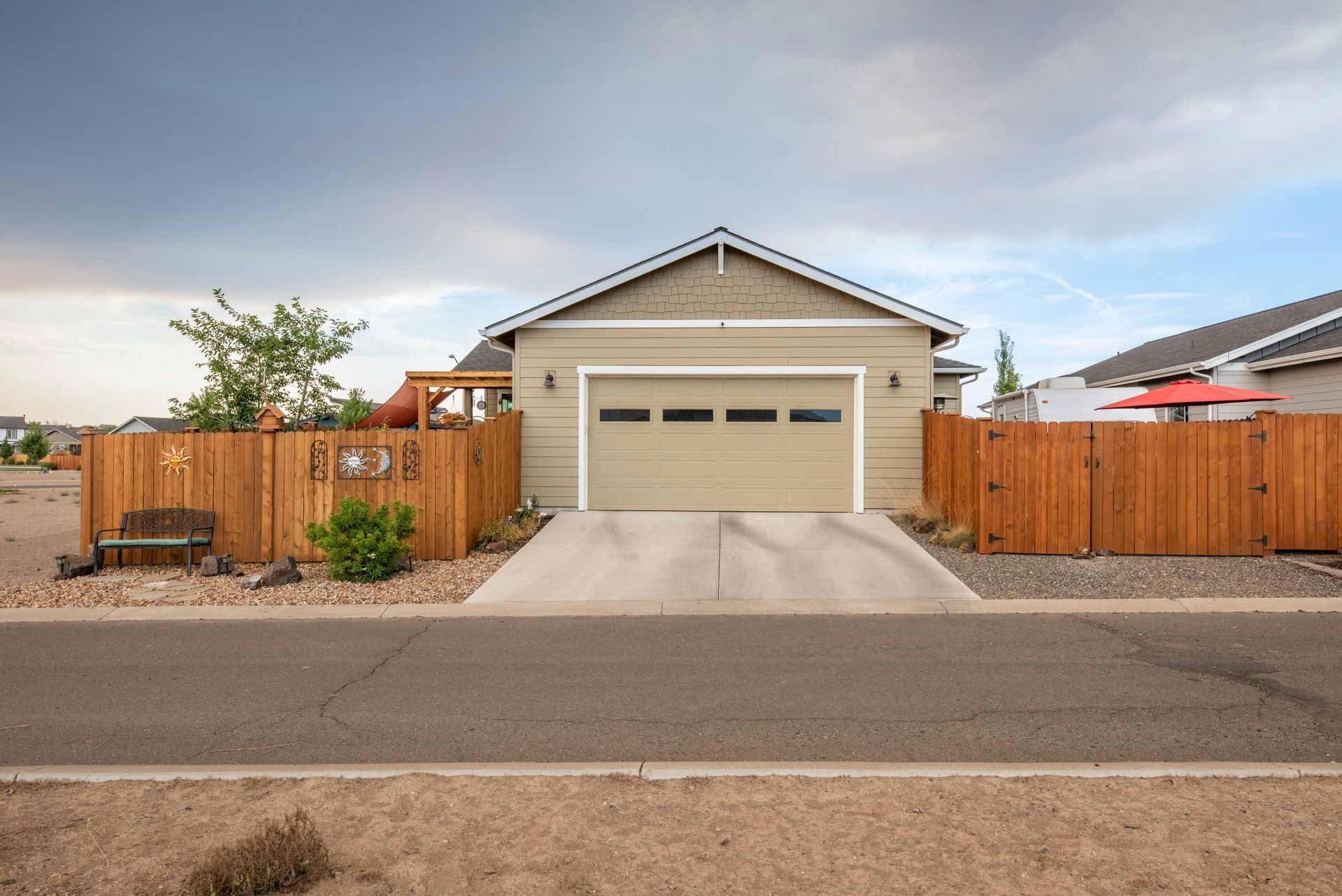 A beige detached garage with a white door and wood privacy fencing, situated behind an asphalt street under a cloudy sky.