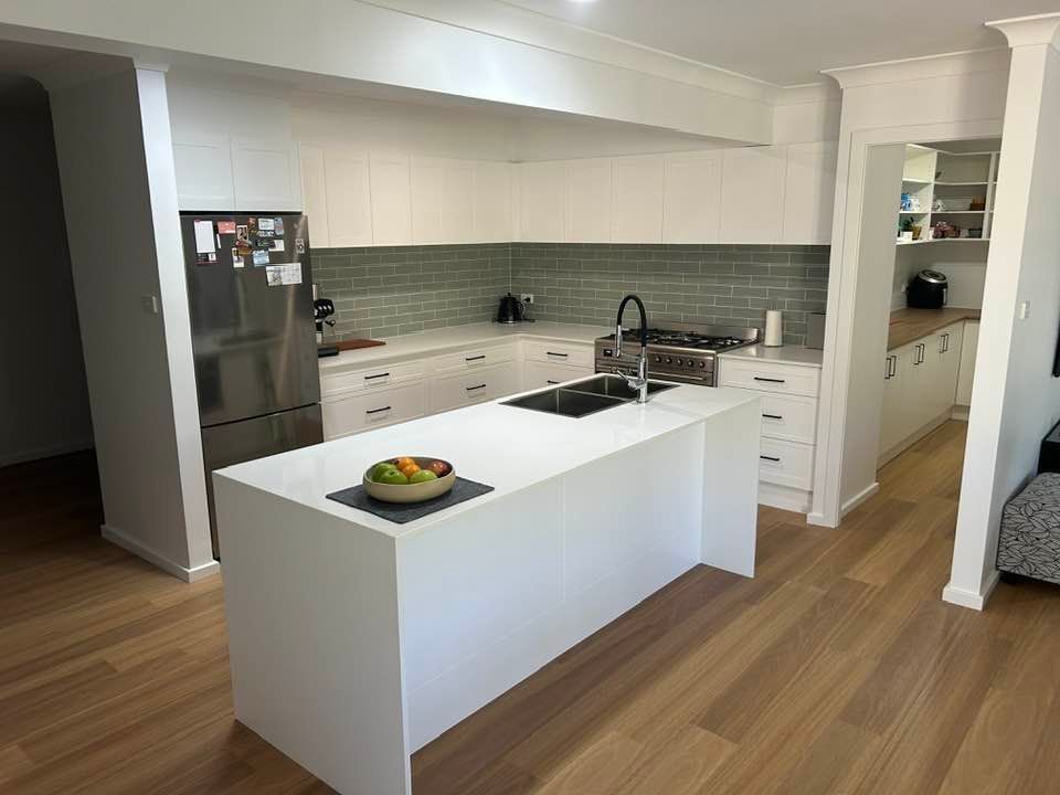 A modern white kitchen featuring a large island, stainless steel appliances, light wood flooring, and green tile backsplash.