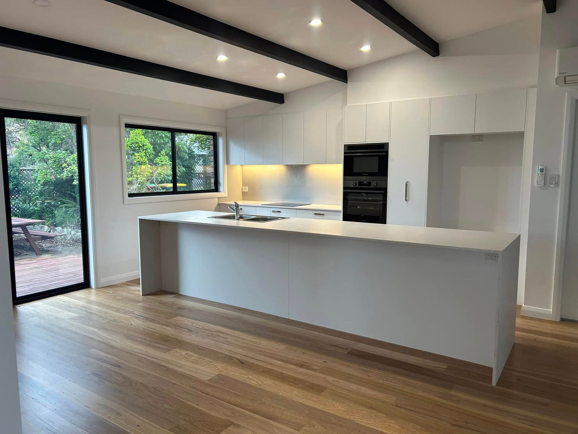 A modern kitchen with white cabinets, a large island, light wood flooring, dark ceiling beams, and a view of a garden.
